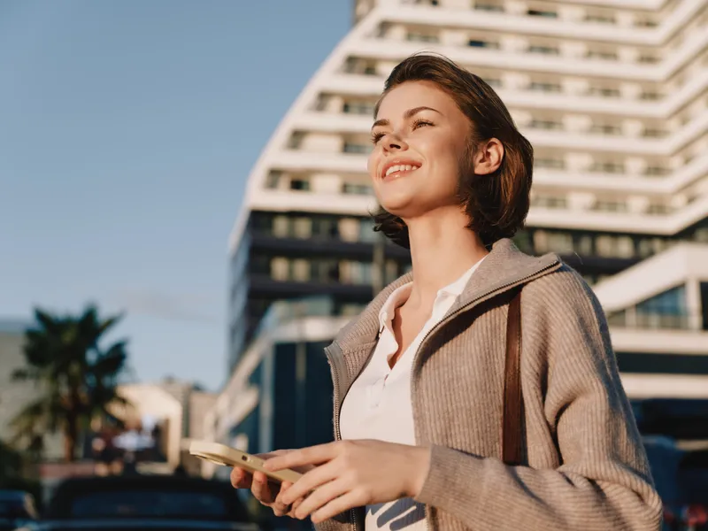 A cheerful woman in business attire stands outdoors, smiling while looking at a smartphone, in a modern urban setting that exudes confidence and positive energy.