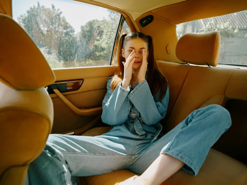 Young woman expressing emotions in a car interior, wearing denim outfit, vibrant background with natural light and warm tones, casual and relaxed vibe.