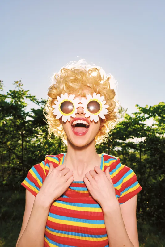Joyful woman in outdoor setting wearing a striped rainbow shirt and sunflower sunglasses, laughing brightly, capturing a playful retro film color lifestyle moment under a clear blue sky