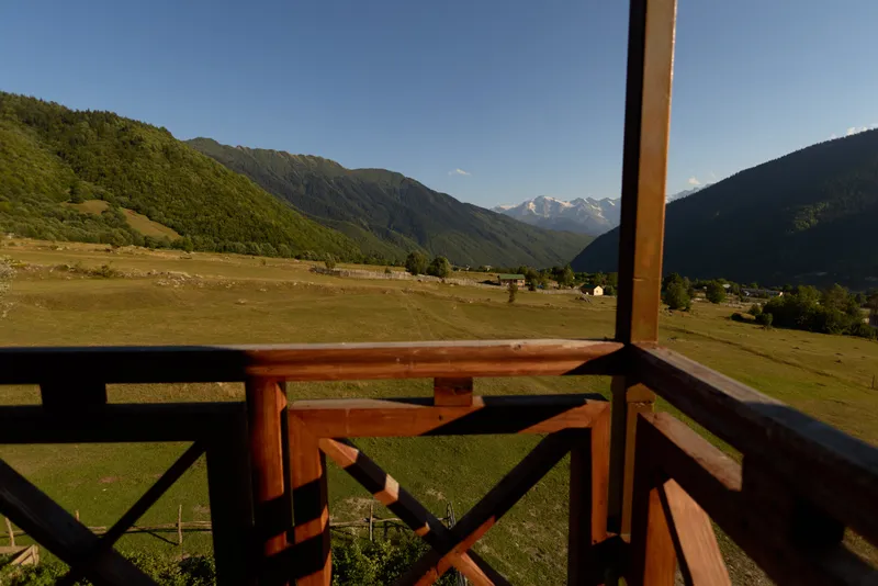 Balcony wooden railing overlooking mountains and valley with green meadow and countryside field, rustic cabin view under clear sky and distant snowy peaks for nature retreat.
