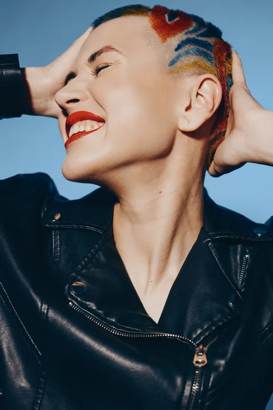 Smiling woman with colorful shaved hairstyle and red lipstick wearing black leather jacket expressing joy and confidence against clear blue sky background.