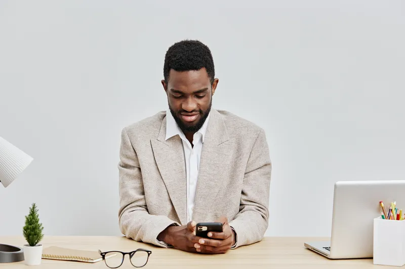 Professional African American man wearing beige blazer and white shirt sitting at desk, using smartphone, with laptop and eyeglasses on table, modern office environment, business work, technology, com