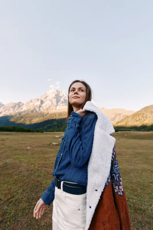Woman in coat over shoulder standing in mountain meadow, portrait looking up with confident smile. Outdoor travel fashion scene in scenic landscape with warm autumn light.