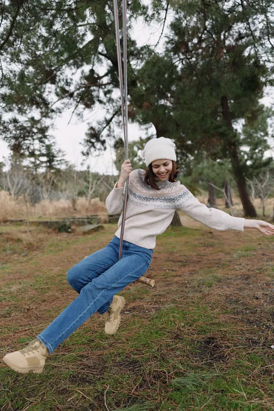 Girl on a wooden swing in an outdoor setting, wearing a knit sweater, beanie and jeans, enjoying a playful, carefree moment among trees and open natural space.