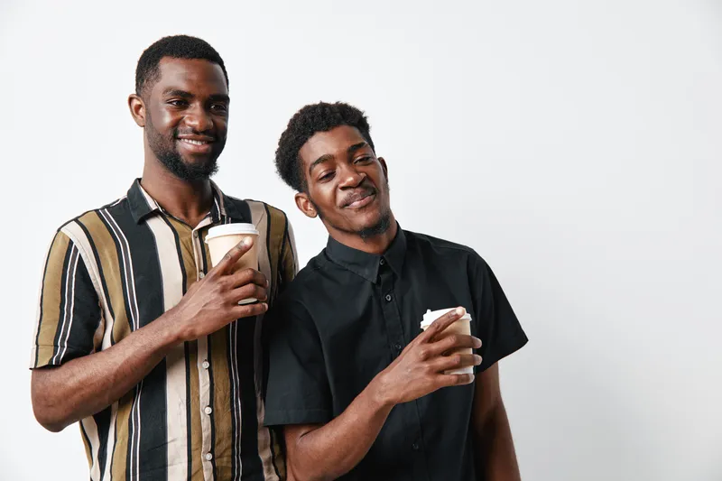 Two young black men with dark skin smiling and holding disposable coffee cups standing against white background. Casual clothing, friendship, and relaxed mood in studio setting.