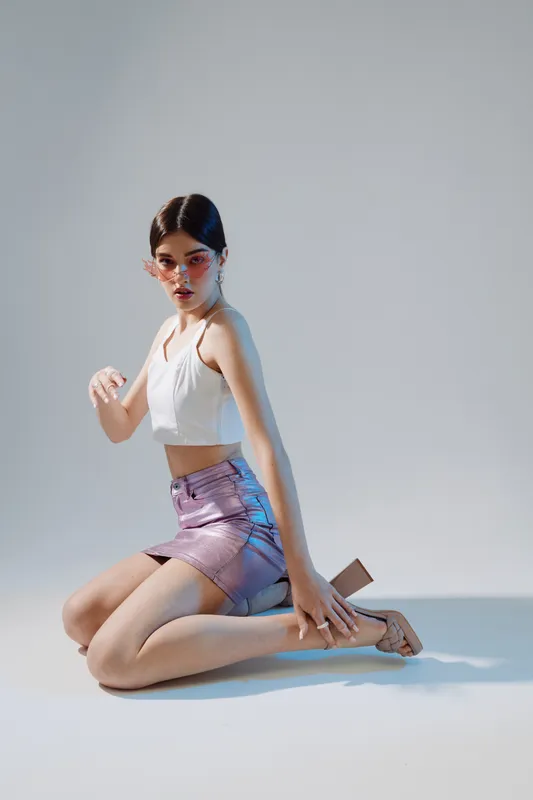 Young woman in a white crop top and shiny pink skirt poses kneeling on the floor against a light background. Concept of beauty, fashion, and modern style with confident expression.
