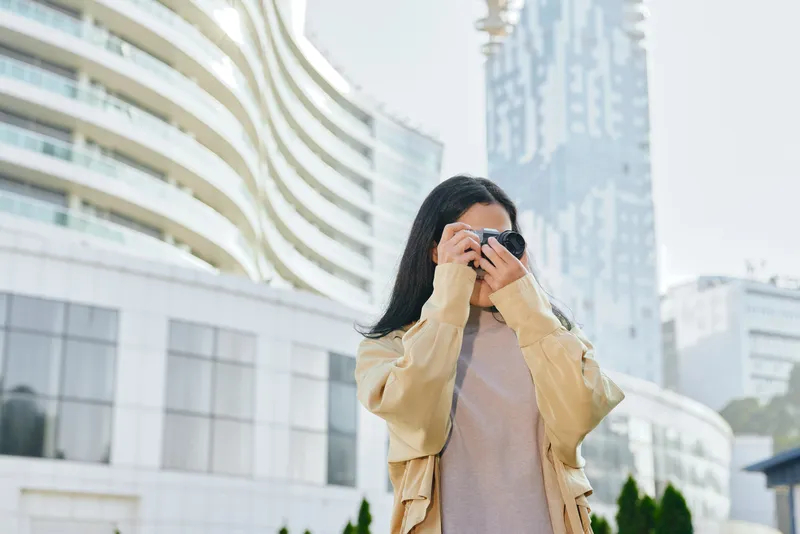 Woman in a beige jacket takes photos with a smartphone in a modern urban setting, glass buildings in the background, candid street photography conveying contemporary city vibes