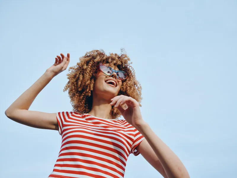 A cheerful figure with curly hair wears a red and white striped shirt and stylish sunglasses, laughing under a clear blue sky, capturing a bright carefree moment.