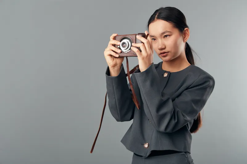 Young woman with long dark hair holding vintage camera, focused on photography. Casual outfit and grey background with creative retro photo concept for hobby or art.