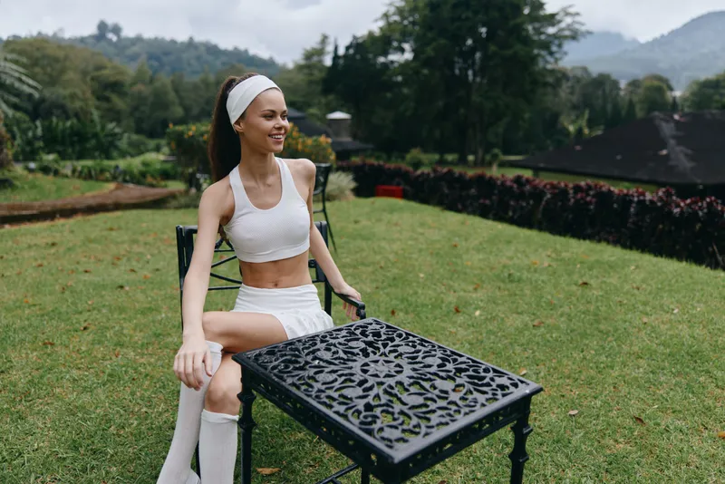 Fit young woman in a white sports outfit sitting outdoors, smiling and enjoying nature, showcasing a healthy lifestyle amidst a vibrant green background