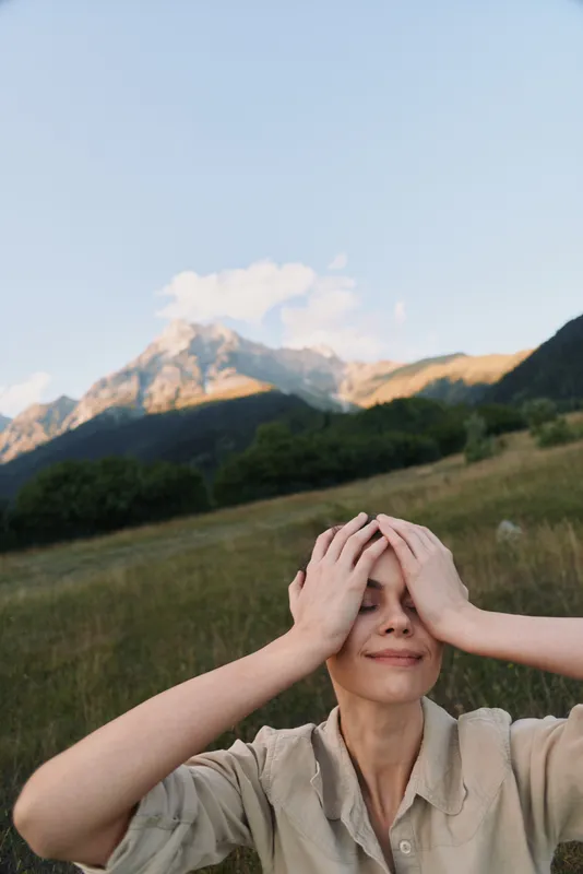 A woman outdoors in a mountain meadow holds her head with both hands, smiling softly. A moment of relief and self care in nature, capturing a lifestyle scene of calm, balance, and personal reflection.