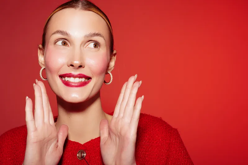 Smiling woman with red lipstick and hoop earrings poses with hands near face on red background. Concept of beauty, fashion, makeup, and confident female portrait.