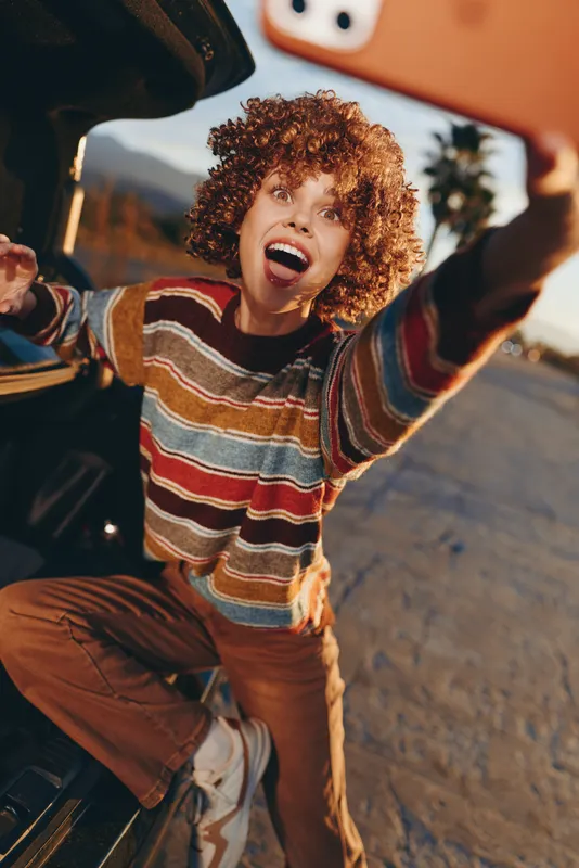 Woman lifestyle scene with curly hair laughing in a car taking a selfie with a phone, striped sweater and jeans, bright sunlight, joyful street travel moment