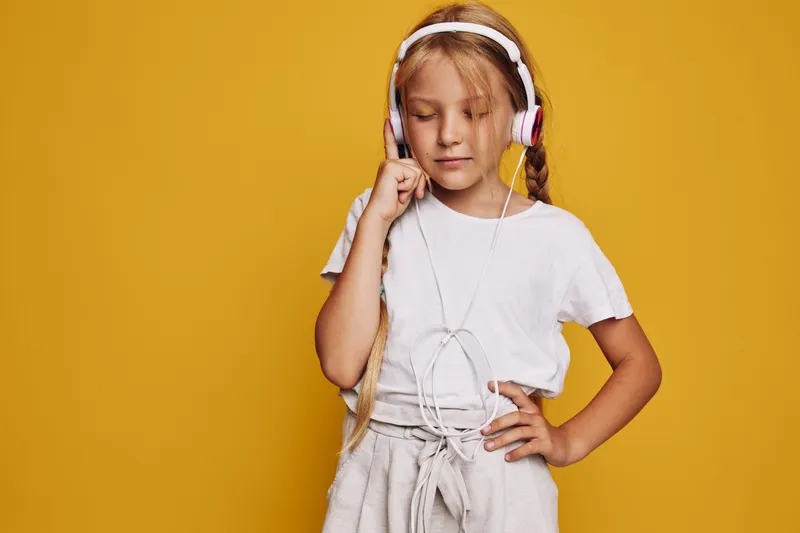 Blonde girl in white t-shirt listens to music with headphones standing against yellow background. Casual style with relaxed expression.