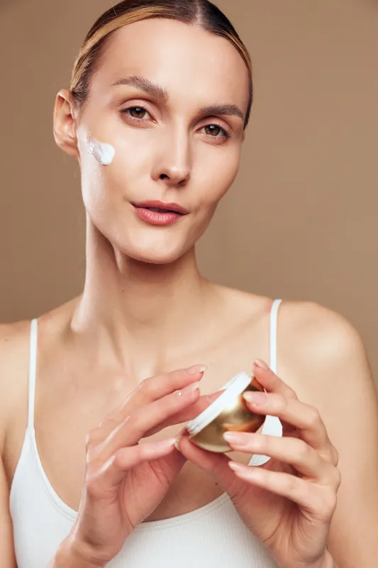 Young woman with clean skin applies moisturizing cream on her face holding cosmetic jar in hand isolated on beige background closeup portrait of skincare beauty routine