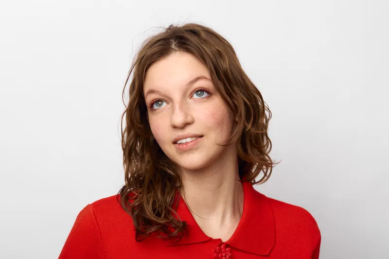 Young woman with curly brown hair wearing red sweater looks up thoughtfully on white background. Concept of contemplation, hope, and natural beauty in casual portrait.