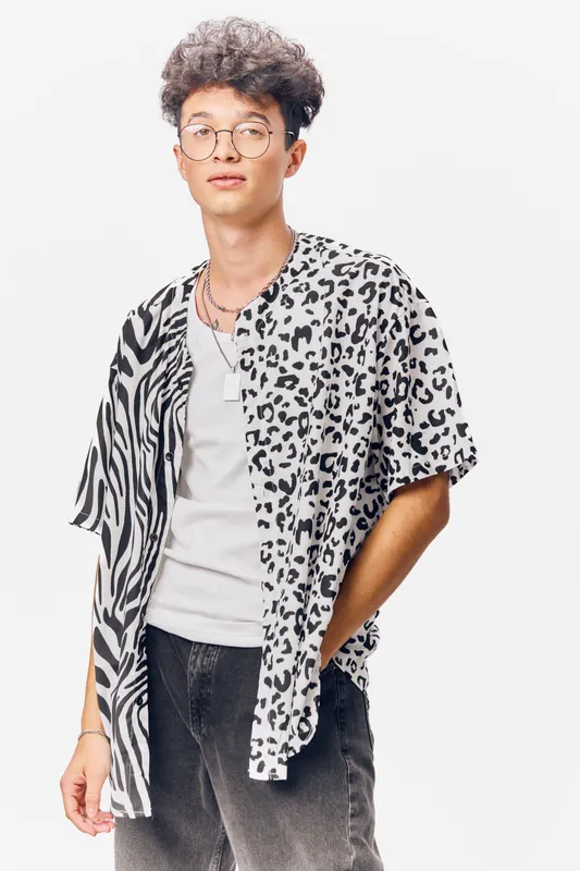 young man with curly hair wearing round glasses and a black and white patterned shirt stands confidently against a white background, showcasing casual street style with relaxed posture
