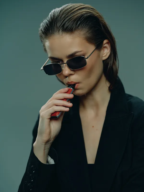Stylish woman vaping with sunglasses wearing black outfit against green background