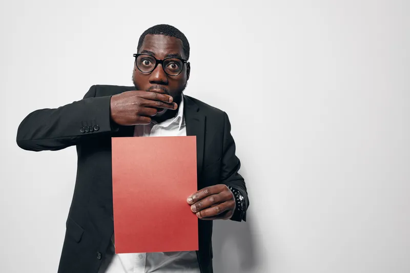 African American man with dark skin and glasses holding red paper covering mouth with hand in surprise or shock. Business casual attire with white shirt and black jacket on white background.