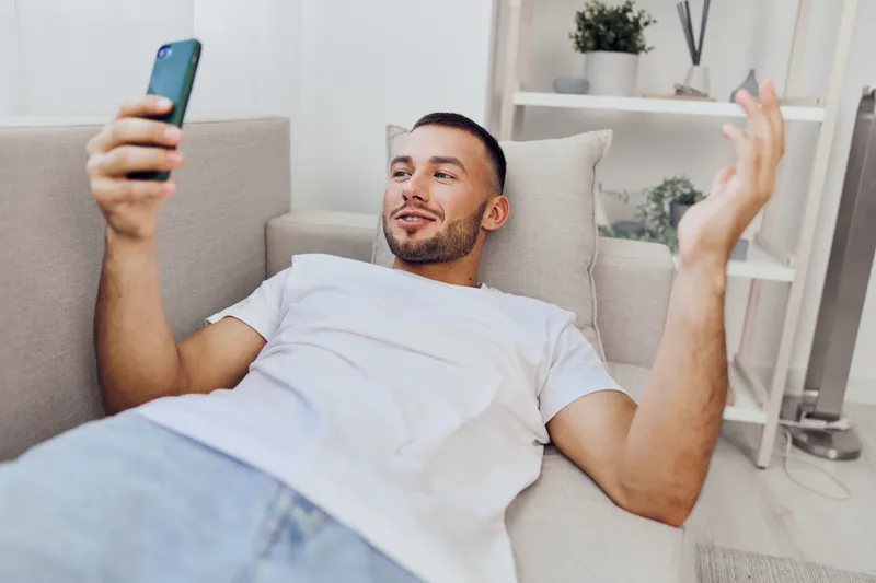 Happy man relaxing on a couch, engaging with his smartphone. Modern home interior with bright colors, capturing the essence of leisure and technology in 2025.