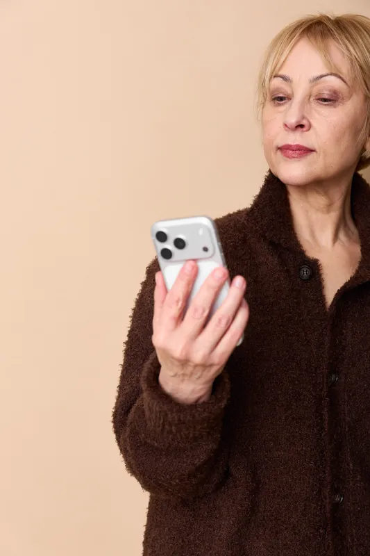 Senior woman holds a smartphone indoors, thoughtful expression, hands around the device, concept of communication, technology, portrait about aging and online.