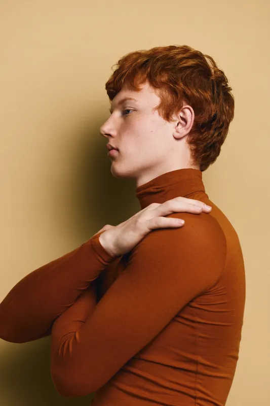 Young man with red hair in brown turtleneck sweater poses in profile against beige background. Casual portrait with folded hands and thoughtful expression for fashion or lifestyle.