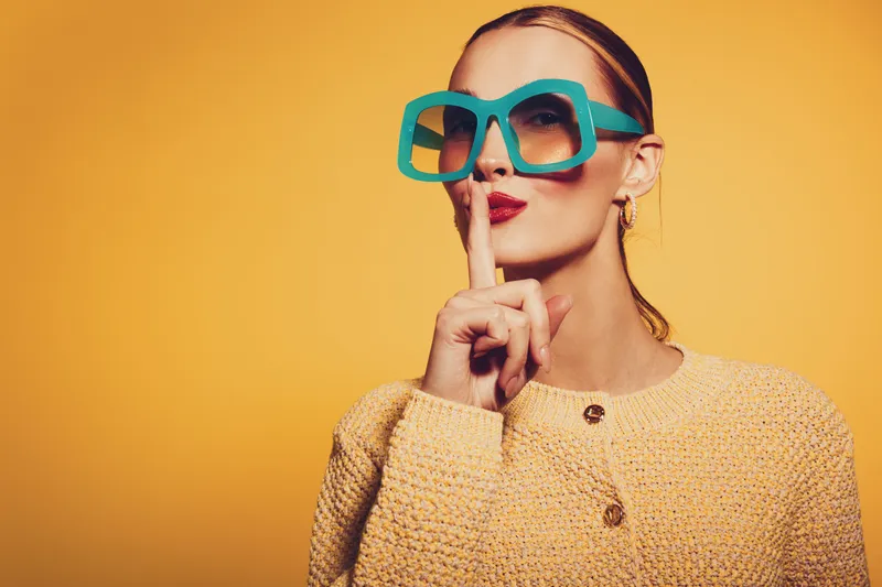 Stylish woman in large blue sunglasses and yellow sweater makes shushing gesture with finger on lips. Bright orange background highlights fashion and secrecy concept in studio portrait.