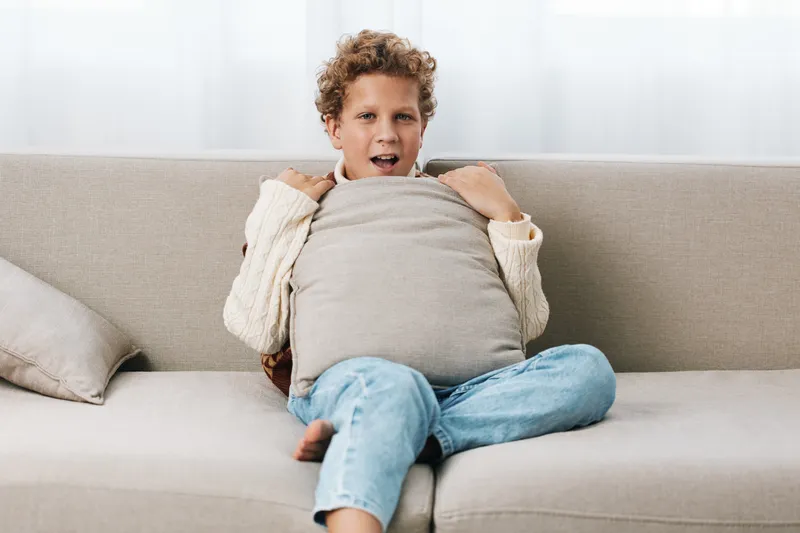 Joyful child sits on a light sofa, hugging a pillow and peeking over it with a bright smile. Cozy sweater and jeans create a warm, homey mood.