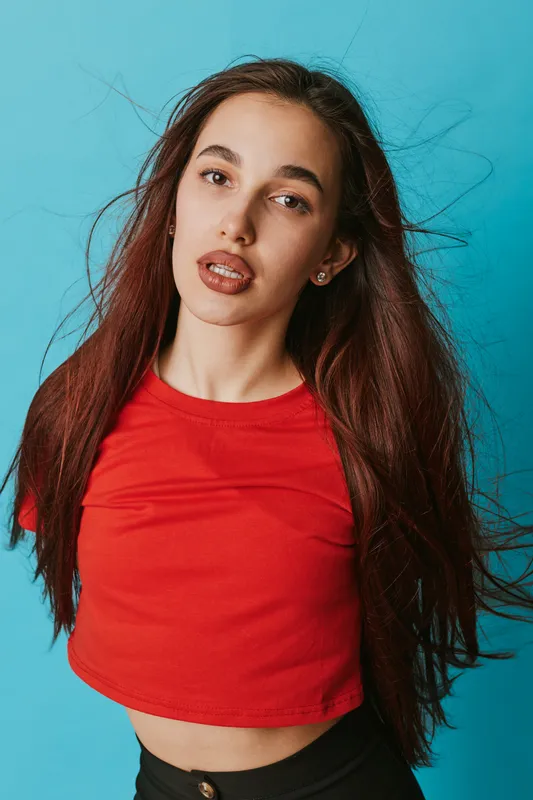 Young woman with long hair wearing red crop top posing against blue background. Casual style, natural makeup, and confident expression for fashion or portrait photography concept.