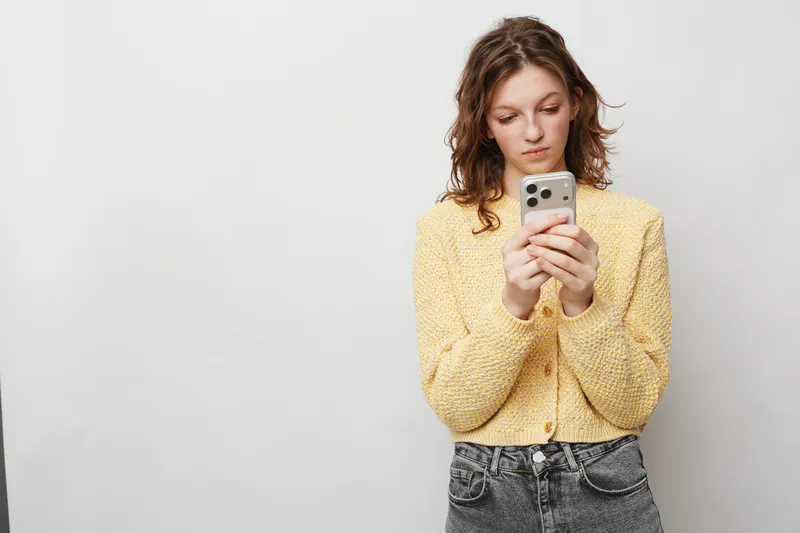 Young woman in yellow sweater looks at smartphone with focused expression. Casual style, indoor studio with plain background. Concept of communication, technology, and social media use.