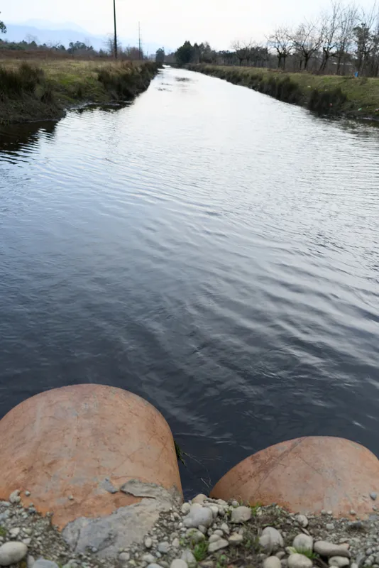 Wastewater discharge pipe on a river bank, calm rippling water in a rural outdoors landscape, overcast daylight, nature and outdoor lifestyle, environmental concern