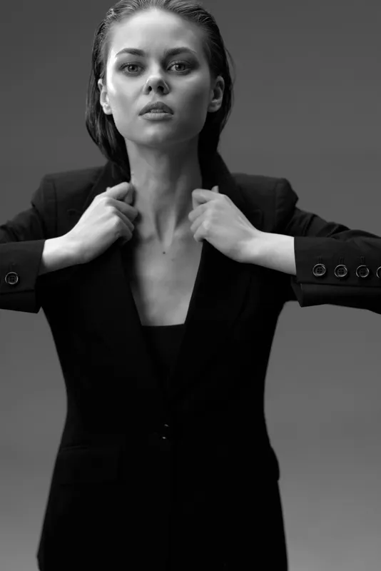 Black and white portrait of confident woman adjusting blazer with serious expression in studio lighting.