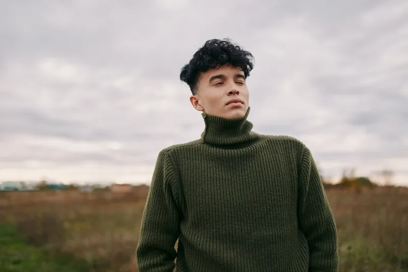 A candid portrait of a young man in an olive green knit turtleneck, capturing authenticity through natural light and a restrained, open field setting.