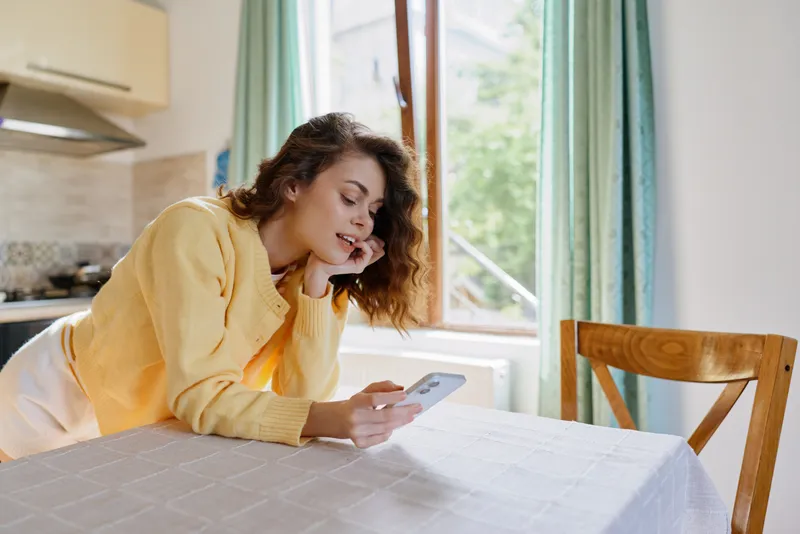 Relaxed woman in a yellow cardigan sits at a table in a sunlit kitchen, looking at a smartphone. Casual home lifestyle, daytime setting, soft natural light, candid moment of everyday comfort.
