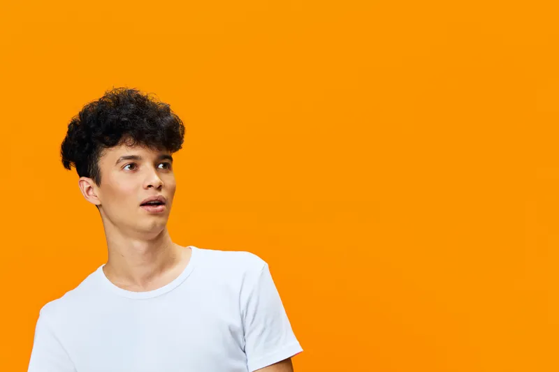 young man with curly hair wearing white t-shirt looking surprised on vibrant orange background, expressive facial expression, casual style and bright contrast