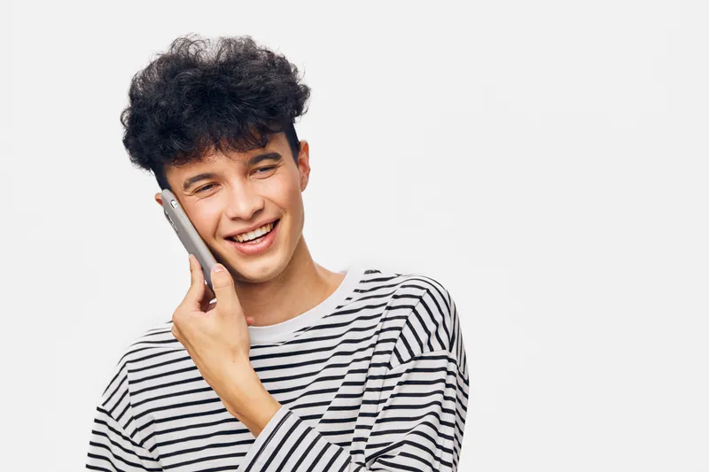 smiling man talking on smartphone wearing striped shirt with curly hair against neutral background, casual communication and modern technology concept