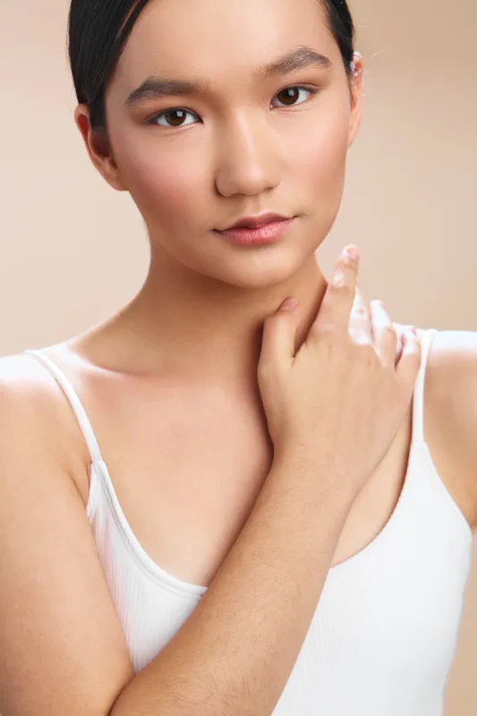 Asian woman appears in a clean studio portrait wearing a white tank top. She gazes softly at the camera with a calm, confident expression, highlighting natural beauty.