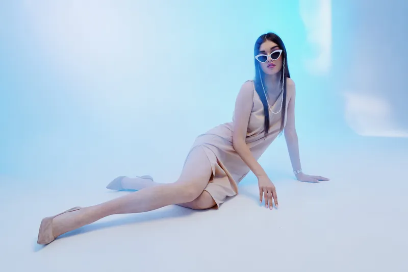 Young woman in a stylish beige dress and white sunglasses poses on the floor in a blue studio. Concept of woman beauty fashion, elegance, and modern style.