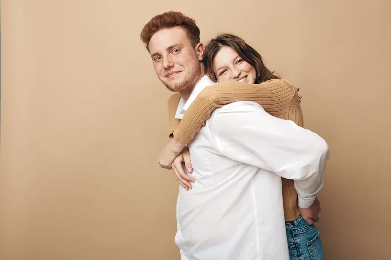 Happy young couple in casual clothes smiling and embracing each other against beige background. Concept of love, friendship, relationship, and positive emotions.