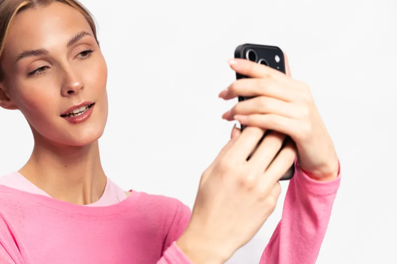 Young woman in pink sweater takes selfie with smartphone on white background. She smiles slightly, capturing a candid moment focused on digital lifestyle and social media use.