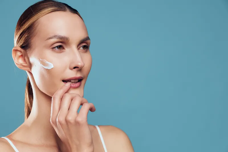 Woman applying skincare cream to cheek, beauty portrait on blue background, gentle finger touch, calm confident expression, smooth face, natural light spa wellness mood