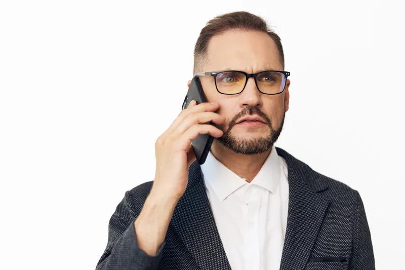 A professional businessman speaks on a mobile phone against a clean colored background, showing a focused expression and confident stance. This isolated portrait conveys urgency and proactive