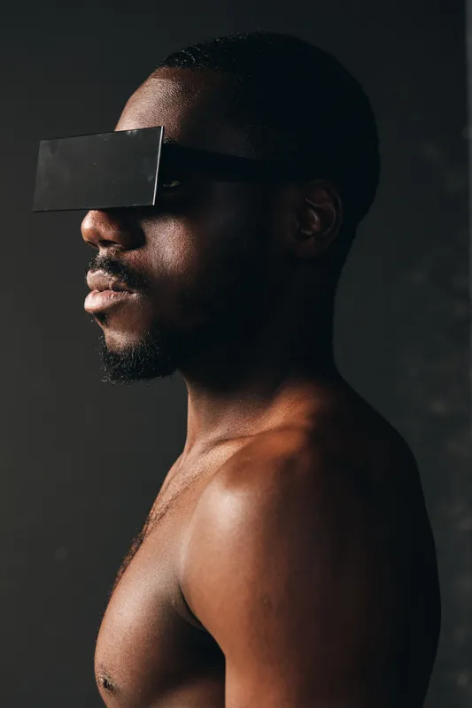 Creative conceptual portrait of African man with dark skin wearing futuristic black visor glasses in studio. Visual style emphasizes modern technology and unique fashion in closeup profile view.
