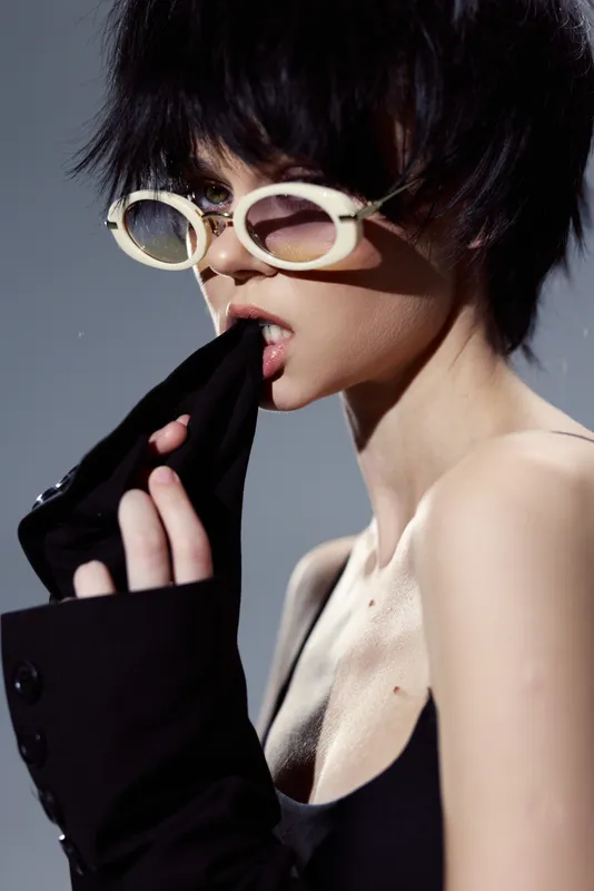 Stylish woman with short hair wearing round sunglasses biting black sleeve, modern fashion, edgy look, confident attitude