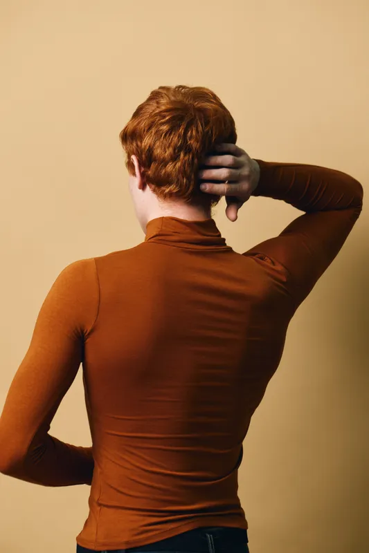 Person with red hair wearing brown long sleeve shirt standing back to camera against beige background touching neck with hand. Casual fashion and portrait style image.