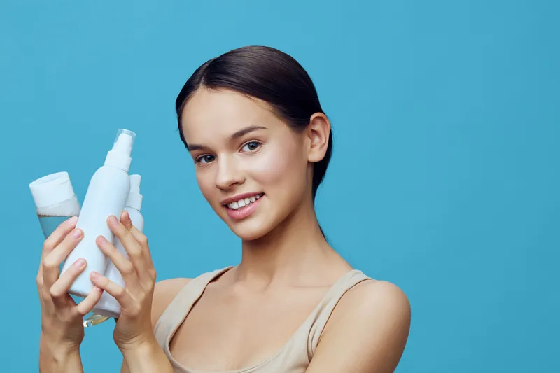 Young woman holding skincare products with a smile against a blue background, representing beauty and self care in a modern aesthetic
