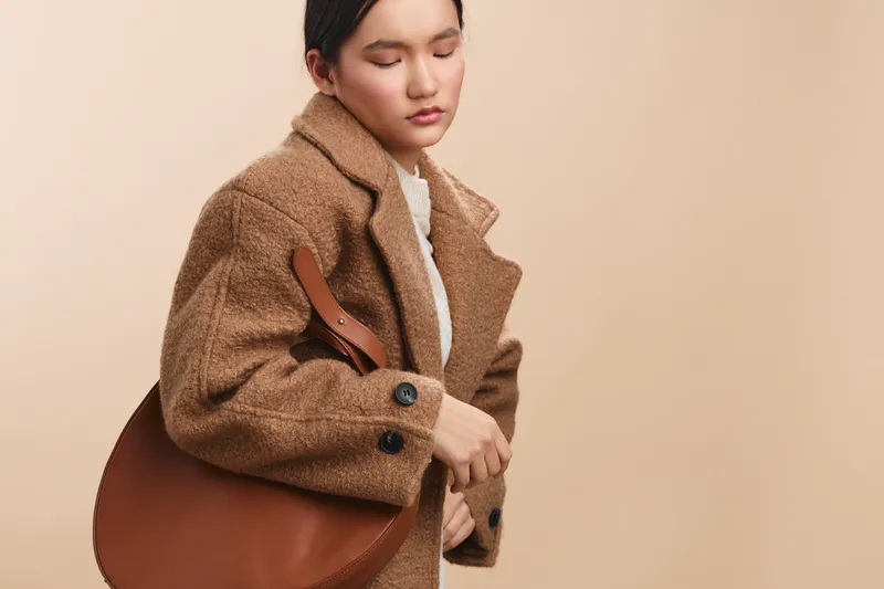 asian woman wearing a brown coat with a leather bag over the shoulder standing against a neutral backdrop, presenting contemporary fashion with poise and quiet confidence.