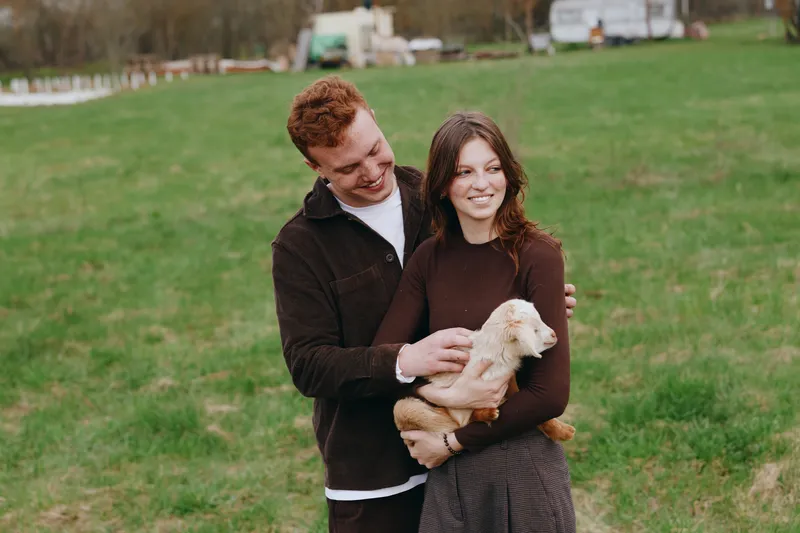A young couple enjoys a moment outdoors as the woman holds a baby goat while the man embraces her from behind in a casual, green field setting during a lifestyle shoot.