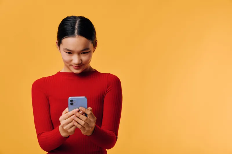 Young woman wearing red sweater looking at smartphone with smile on orange background. Asian girl holding phone using social media, texting message and browsing mobile device.