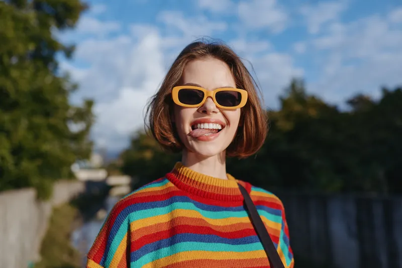 Young woman in a rainbow sweater and yellow sunglasses smiling playfully, short bob haircut, casual outfit outdoors by an urban canal in sunny daytime, lifestyle travel portrait.